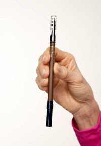 Crayon à sourcils marron avec un design à torsion ; comporte un capuchon transparent et une petite brosse à l'extrémité opposée. Main tenant le crayon.