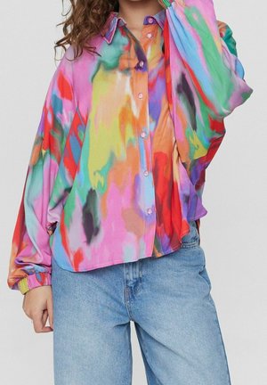 Persoon draagt een loszittend button-downoverhemd met lange mouwen in multicolor tie-dye, gecombineerd met lichtblauwe spijkerbroek, poseert met één hand nabij het gezicht.