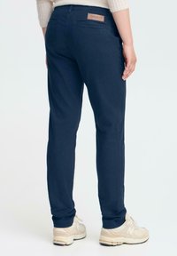 Oseba v temno modrih hlačah in svetlih bež športnih čevljih stoji ob ozadju brez vzorcev, prikazana s hrbtne strani.