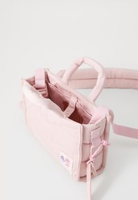 Sac en nylon rose doux avec une structure rembourrée, de courtes poignées et une bandoulière réglable. Présente un écusson floral et une couture décorative.