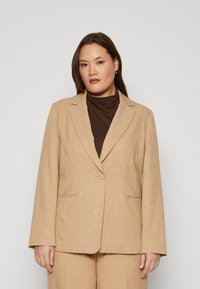 Beige blazer i ett strukturerat tyg med enkel knappstängning, slagkrage och två framfickor. Bärs över en brun polotröja.