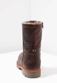 Bottine en cuir marron avec une doublure douillette, bout arrondi et semelle en caoutchouc basse. Présente un accent logo en métal près du talon et des coutures verticales.
