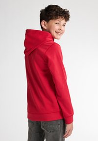 Felpa rossa con cappuccio, dal texture morbido, con polsini a costine e tasca a marsupio. Indossata sopra jeans grigi, mostra un taglio casual.