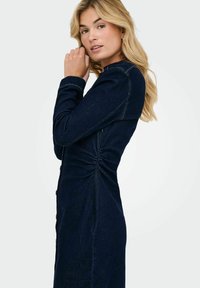 Robe en denim foncé avec boutonnage à l'avant et détails froncés à la taille. Elle présente des manches longues et une silhouette ajustée.