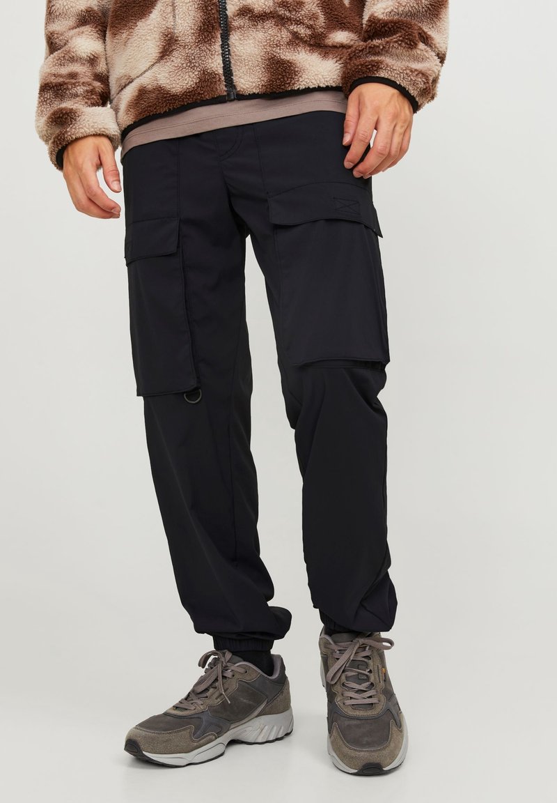 Černé cargo kalhoty s elastickými manžetami, několika bočními kapsami a hladkou texturou, v kombinaci se šedými teniskami. Fleece bunda má hnědý vzor.