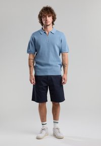 Light blue short-sleeve polo with a zip collar, textured knit fabric, paired with dark navy shorts and white sneakers with green-striped socks.