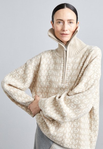 Beige zippered tröja med ribbad krage och breda ärmar, som har ett subtilt mönster av sammanflätade bokstäver i en mjuk stickad textur.