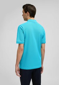 Turquoise poloshirt met een effen textuur, korte mouwen en een geknoopte halslijn, van achteren weergegeven. Gecombineerd met donkere broek.