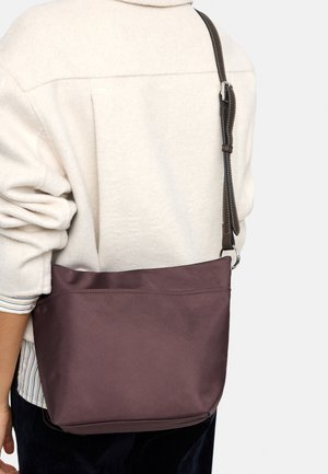 Osoba w kremowej polarowej kurtce, niosąca ciemnobrązową torbę na ramię z regulowanym paskiem i srebrną klamrą.