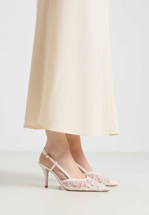 Piedi che indossano décolleté slingback beige in pizzo con punta appuntita e cinturini alla caviglia, abbinati a una gonna crema alla caviglia.