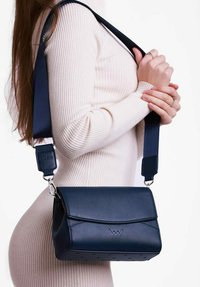 Tmavě modrá kožená crossbody taška s klopou, stříbrným kováním a nastavitelným popruhem. Obsahuje logo a perforované detaily ve spodní části.