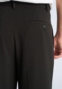 Nærbillede af sorte kjolebukser med en baglomme med knap, båret af en person i en hvid skjorte.