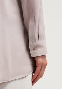 Poignet de manche d'une chemise beige clair avec un bouton, portée par une personne ayant la main détendue le long du corps sur un fond uni.