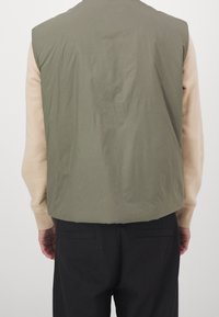 Olivově zelená vesta bez rukávů s hladkou texturou. Má kulatý výstřih a je nošena přes béžové tričko s dlouhým rukávem.