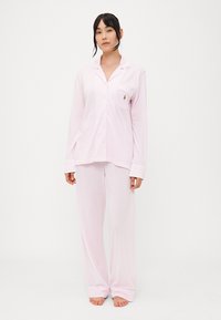 Ροζ βαμβακερό σετ πυτζάμας που περιλαμβάνει ένα κουμπωτό τοπ με μακριά μανίκια και τσέπη στο στήθος, καθώς και αντίστοιχες φαρδιές παντελόνες. Μαλακή υφή, άνετη εφαρμογή.