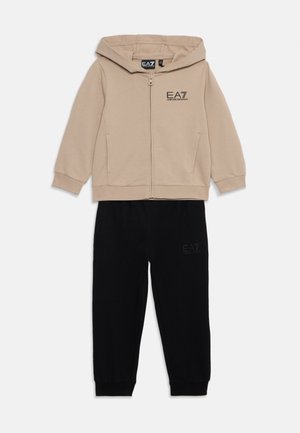 Veste à capuche zippée en coton beige avec poches frontales et logo noir, accompagnée d'un pantalon de survêtement en coton noir avec des détails de logo près de la taille.