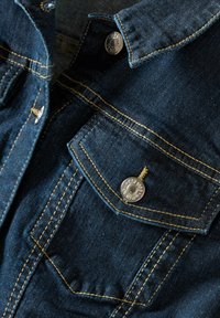 Tummansininen farkkutakki, jossa on terävä kaulus, hopeinen metallinappi sekä erottuva keltainen ommel rintataskussa ja saumoissa.