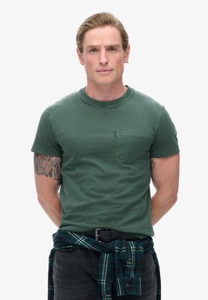 Green cotton T-shirt with a crew neckline and short sleeves, featuring a chest pocket. Black jeans and a blue plaid shirt tied at the waist.