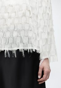 Λευκό υφασμάτινο ύφασμα με παραλληλόγραμμα ανοίγματα και κρόσσια στο μανίκι και το τελείωμα, εμφανίζεται στο χέρι ενός ατόμου με απλό φόντο.