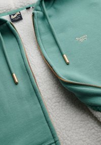 Sudadera verde con cremallera dorada, forro interior suave y logo "Super Dry" bordado en blanco.