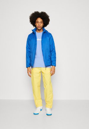 Blauwe lichtgewicht jas met capuchon, gedragen over een lichtblauwe t-shirt. Gele broek en witte sneakers met blauwe accenten maken de outfit compleet.