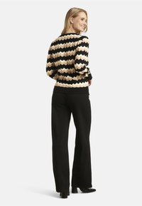 Knitted sweater featuring a chevron pattern in black, beige, and cream, with a ribbed neckline and cuffs. Paired with black trousers.