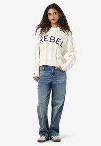 Kremo spalvos megztinis su kabelių raštu, ant krūtinės užrašytas žodis "REBEL" tamsiai mėlyna spalva, derinamas su šviesiai mėlynomis laisvo kirpimo džinsais ir smėlio spalvos sportbatiais.
