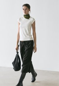 Femme marchant à l'intérieur, portant un haut blanc sans manches, un pantalon large noir, des bottes noires montant jusqu'aux genoux, un foulard vert autour du cou et portant un sac à main noir.