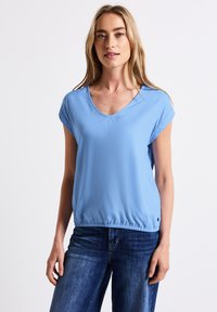 Lichtblauw kortom shirt met een V-hals en elastische tailleband, gemaakt van een zachte, gladde stof. Gecombineerd met donkerblauwe jeans.