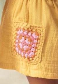 Jupe en tissu jaune avec un patch en crochets texturé présentant un motif de granny square rose et orange le long de l'ourlet.