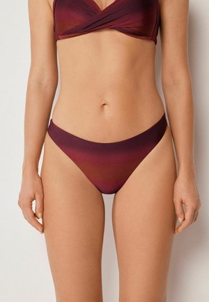 Donna che indossa un bikini color melanzana con top annodato e slip a vita bassa abbinati, in piedi davanti a uno sfondo chiaro e uniforme.