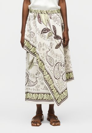 Λευκή μίντι φούστα με πράσινα και καφέ βοτανικά και πεϊσλί σχέδια, συνδυασμένη με καφέ φλατ σανδάλια και λευκό μπλουζάκι μέσα στη φούστα.