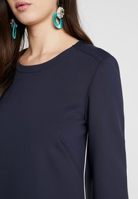 Abito blu navy con una texture liscia, colletto rotondo e dettagli con tasche laterali. Presenta maniche lunghe e cuciture a contrasto bianche.