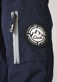 Tissu bleu marine avec une poche zippée dotée d'un curseur noir et un écusson circulaire représentant des montagnes et le logo "Clarks".