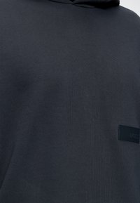 Sweat à capuche gris foncé avec une petite pièce rectangulaire noire présentant un logo subtilement embossé sur la poitrine.