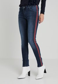 Mörkblå skinny jeans med ett rött och vitt rand ner på sidan, ihop med vita högklackade ankelstövlar. Slät denimtextur.