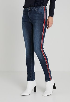 Jeans ajustados de color azul oscuro con una franja roja y blanca en el lateral, combinados con botas de tobillo blancas de tacón alto. Textura de denim suave.