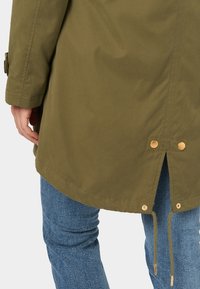 Olivově zelená bunda střední délky s zaobleným lemem, knoflíkovými detaily a nastavitelnými šňůrkami v pase. Hladká textura látky.