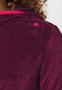 Chaqueta de forro polar en burdeos profundo con una superficie suave y texturizada. Presenta un acento rosa a lo largo del cuello y un pequeño logo bordado en el hombro.
