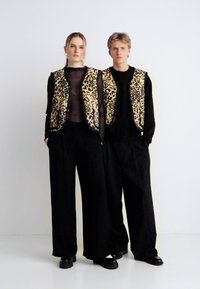 Két modell fekete ruhákat visel, amelyeket leopárdmintás mellényekkel párosítanak. A mellények puha tapintásúak, kontrasztot alkotva a sima, sötét nadrágokkal.