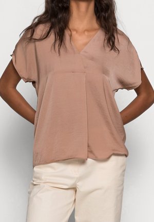 Kobieta ubrana w luźną, krótką bluzkę z dekoltem w kształcie litery V w jasnobrązowym kolorze oraz kremowe spodnie, stojąca ze złożonymi rękami za plecami.