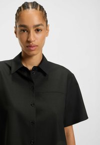 Black short-sleeve shirt with a button-down front, featuring a standard collar, a small chest pocket, and smooth, textured fabric.