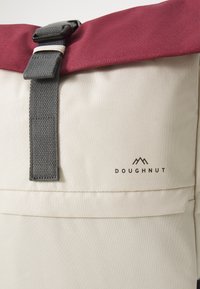 Rucsac din material bej cu un capac maron, curea gri, un buzunar frontal și logo-ul "DOUGHNUT" cu un grafic montan în imprimare închisă.