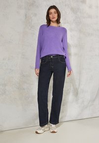 Maglione lavorato a maglia viola con un motivo testurizzato, abbinato a jeans blu scuro e sneakers beige con accenti verdi. Outfit casual caratterizzato dalla semplicità.