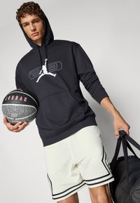 Sort bomulds hættebluse med hvid "Jordan" logo, letvægts hvide mesh shorts med sorte detaljer, holder en mønstrede basketball og en sportstaske.