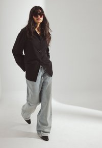 Svart knappad skjorta, ljusblå vida jeans och spetsiga svarta klackar. Modellen bär stora mörka solglasögon. Mjuk belysning, neutral bakgrund.