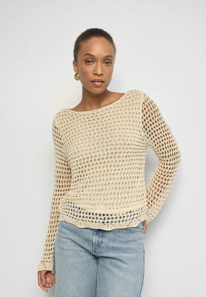 Mujer con suéter beige de punto abierto de manga larga y vaqueros azul claro, de pie frente a un fondo blanco liso.