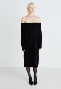 Abito nero fuori spalla con maniche lunghe, realizzato in un morbido tessuto a maglia. Presenta un collo a coste e un taglio dritto, lungo fino alla caviglia.