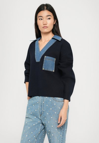 Maglione navy con colletto e tasca in denim, caratterizzato da un design a scollo a V e una vestibilità rilassata. Indossato con pantaloni in denim azzurro chiaro decorati con borchie.