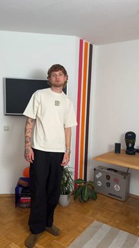 Krémové tričko s krátkým rukávem a malým logem, černé volné kalhoty, světle hnědé boty, viditelné tetování na rukou, jednoduché vnitřní prostředí.
